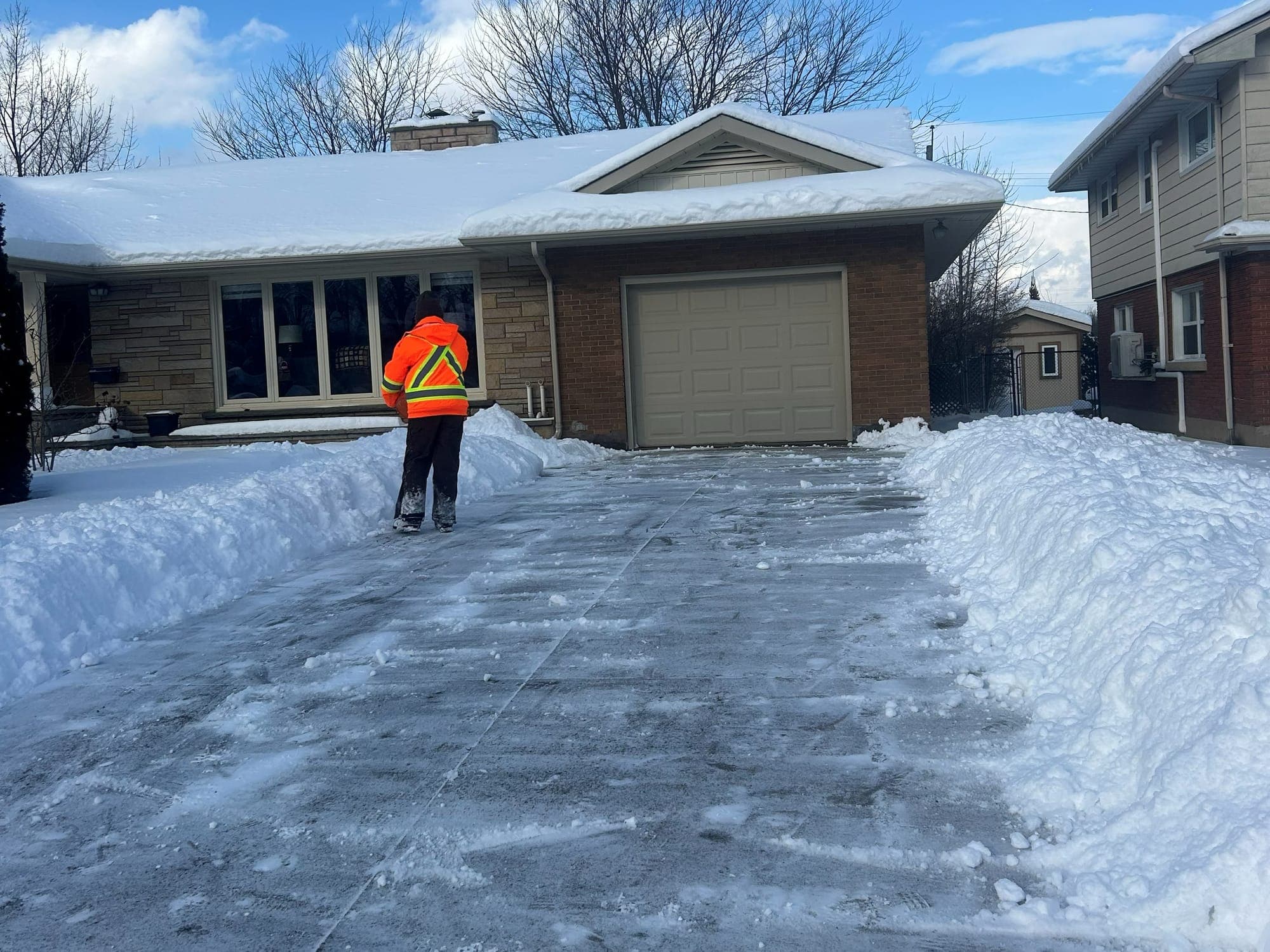 Crew member in a hi-vis orange jacket at the top of a freshly cleared driveway, snow banked high on both sides, in front of a brick and stone bungalow