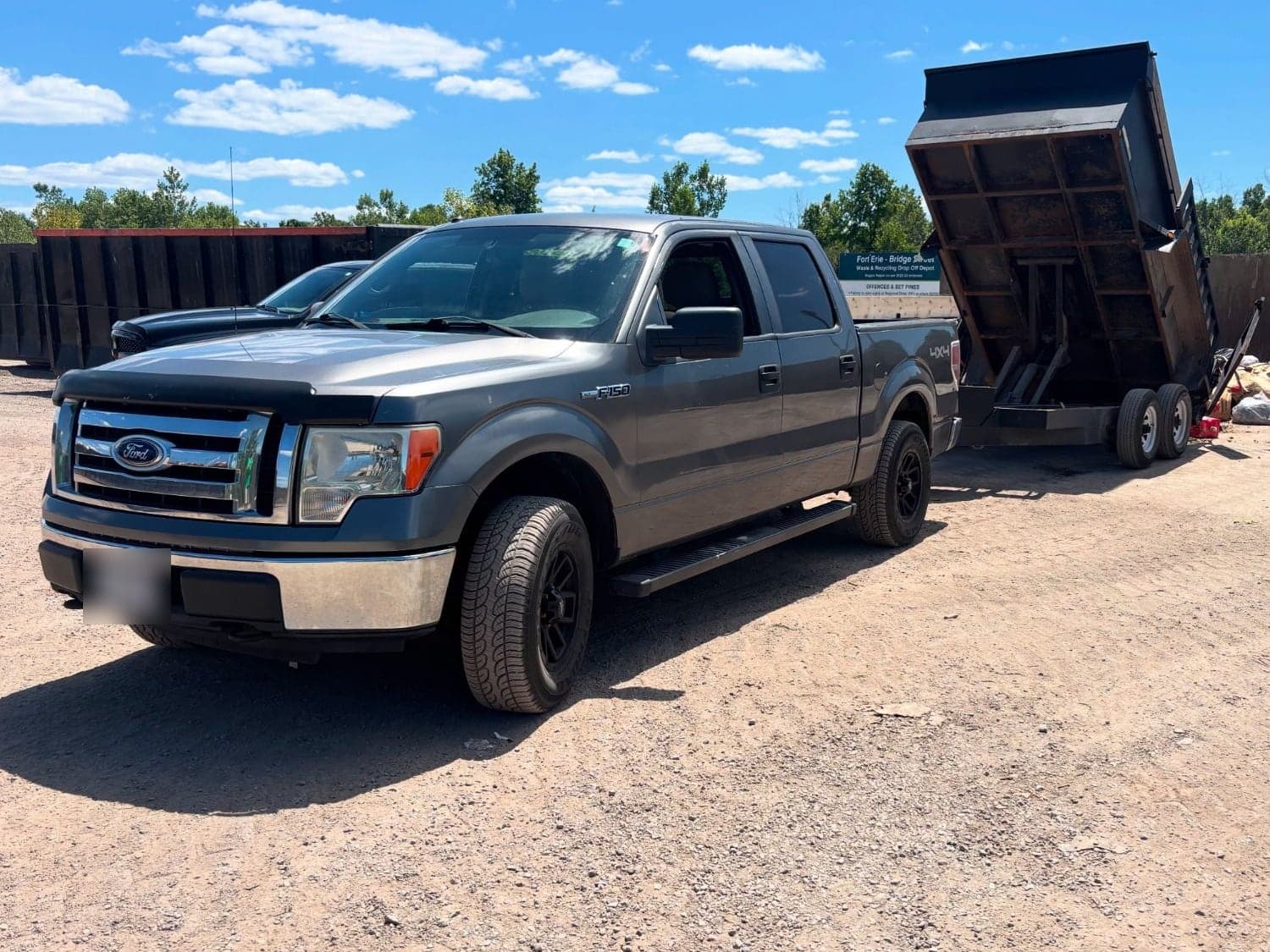 The crew's grey pickup truck hitched to a tipped dump trailer at a regional waste and recycling drop-off depot, after unloading a haul